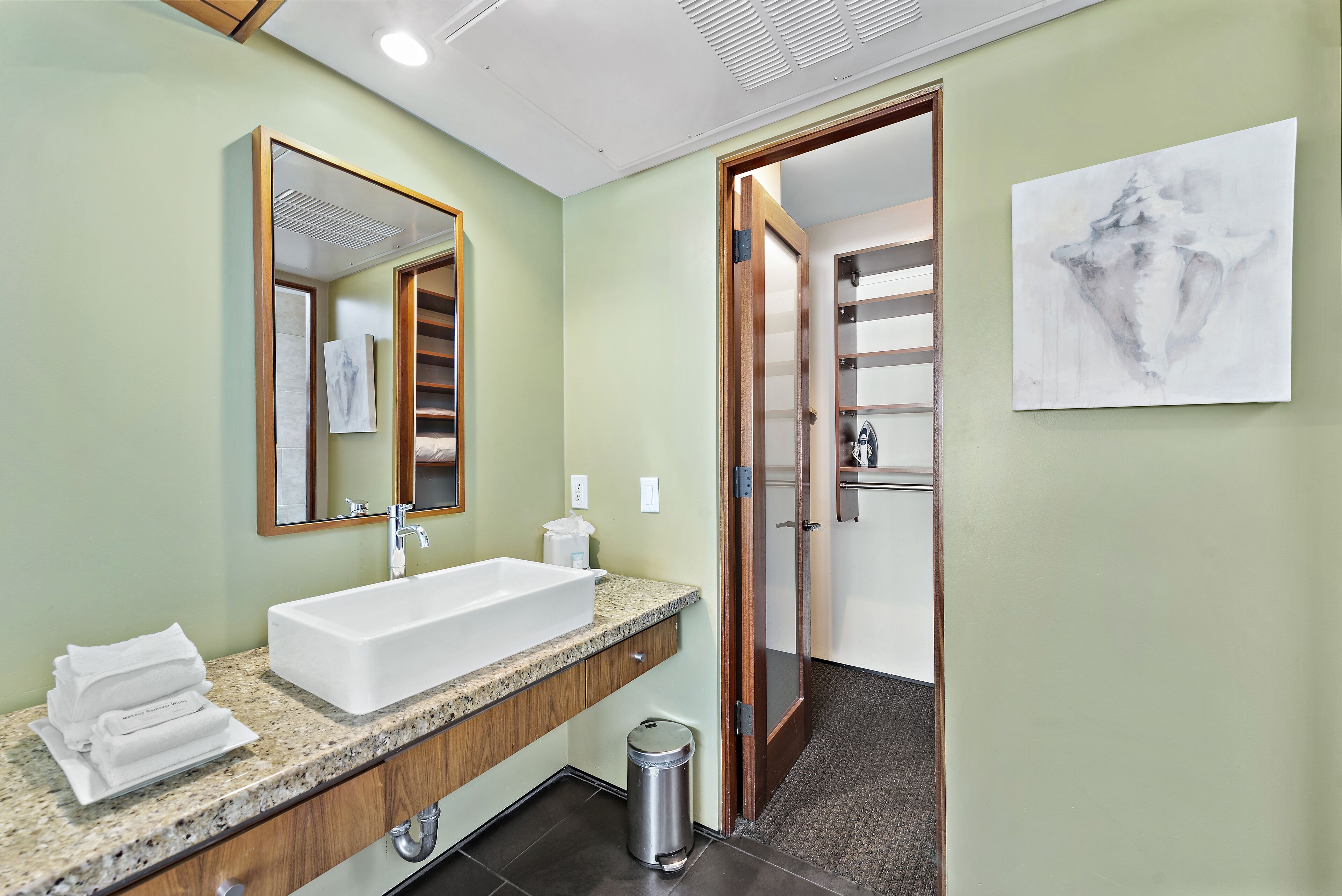 A modern bathroom with a large sink, vanity, and a mirror. There is a trash can and a door leading to a shower area.