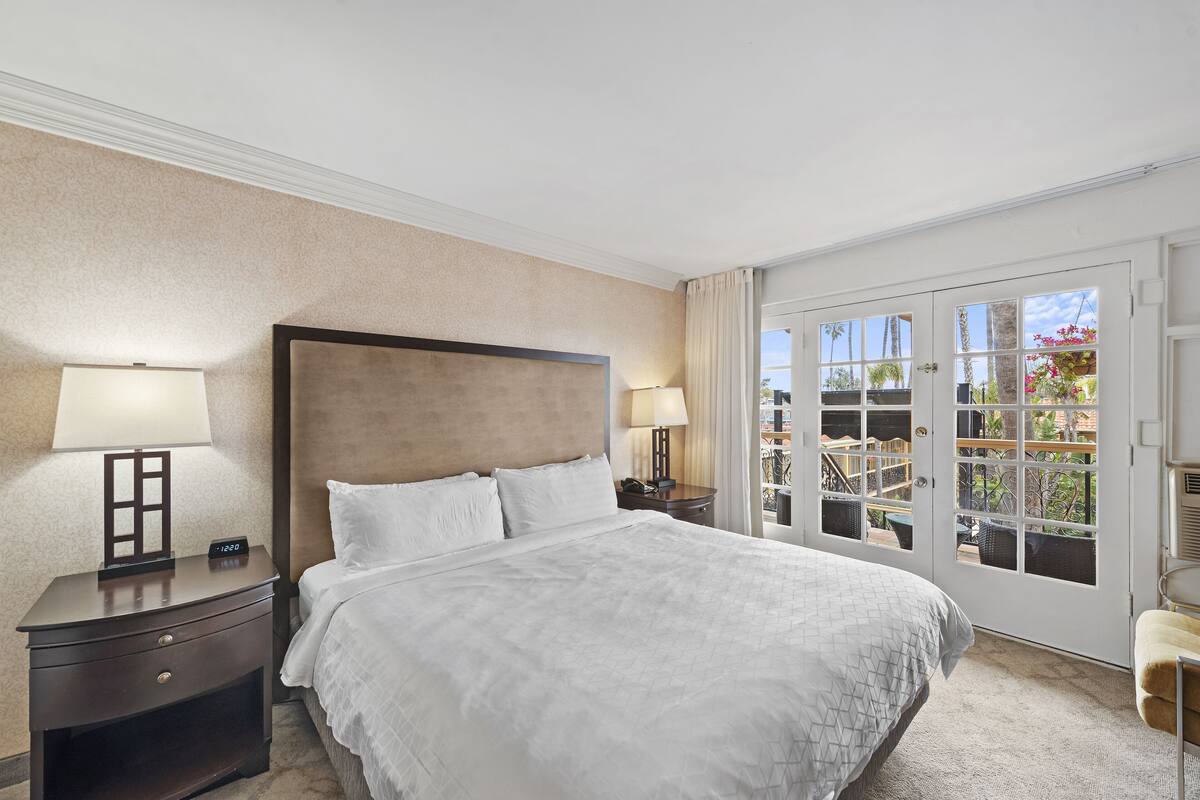 A hotel room with a large bed, two bedside tables with lamps, a view through a glass door to an outdoor area with plants.