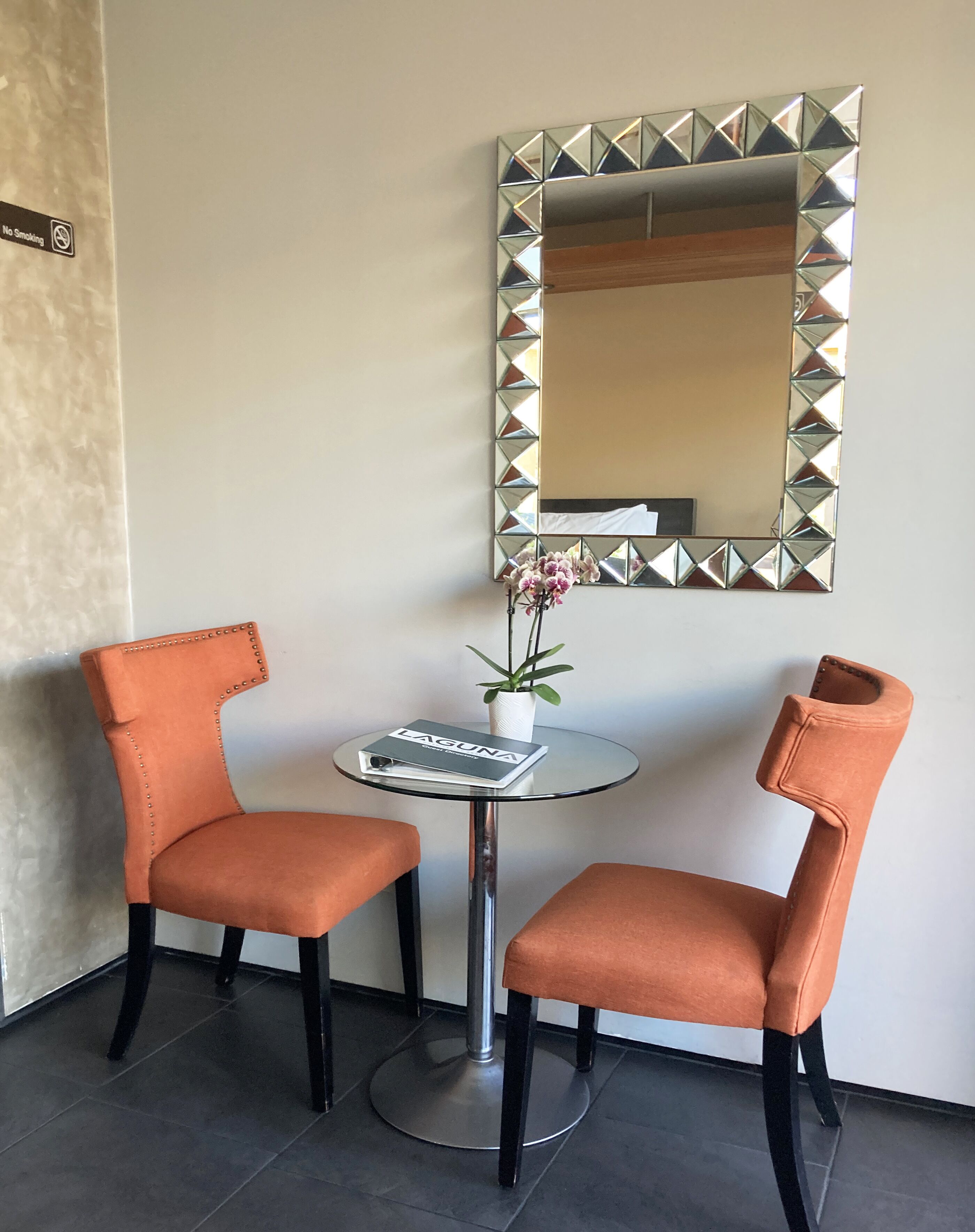 Two orange upholstered chairs with black legs, a small round table with a glass top, a white vase with pink flowers, and a large mirror with a geometric frame on the wall.