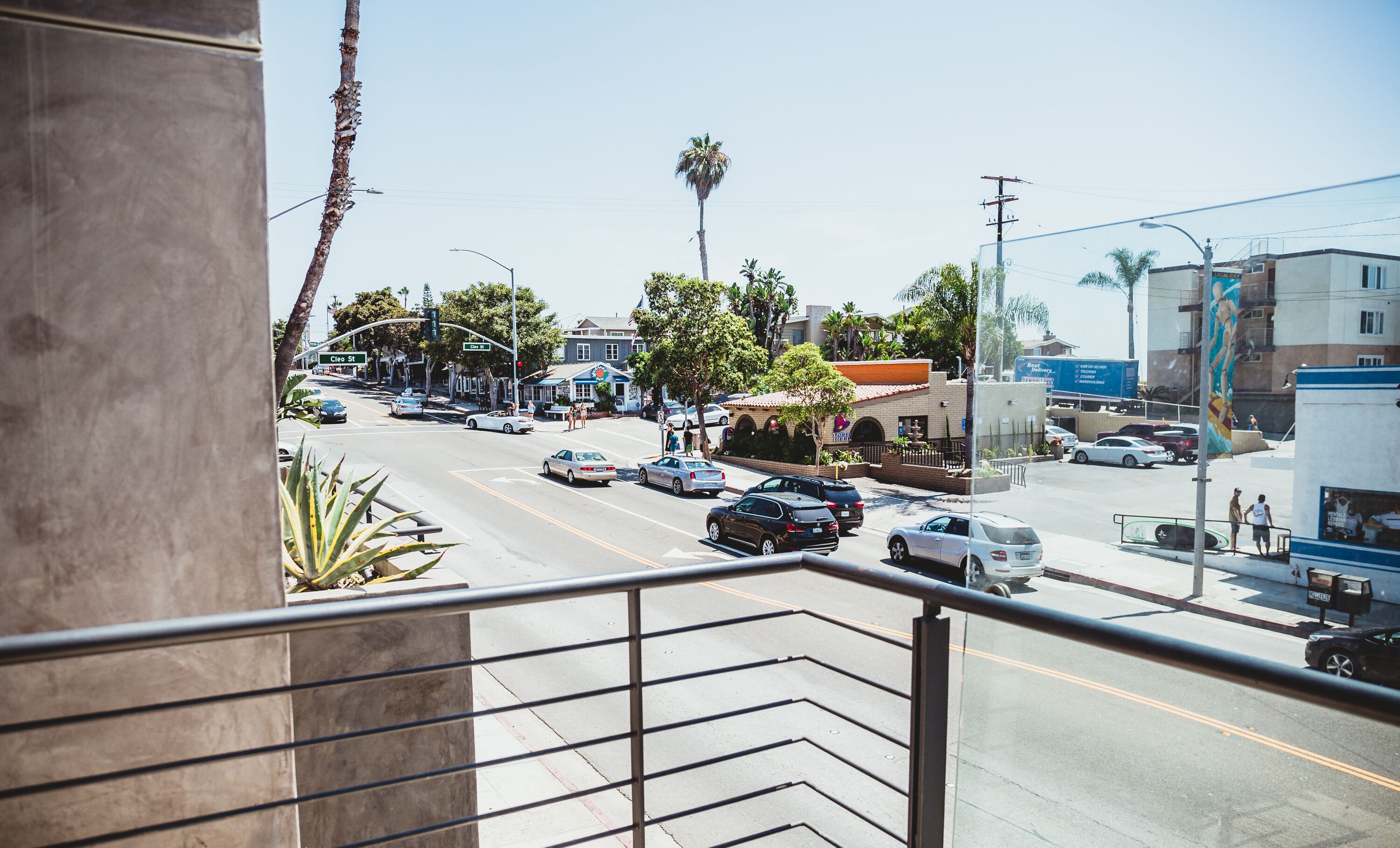 A street view from a balcony with a clear sky, cars on the road, and buildings in the background.