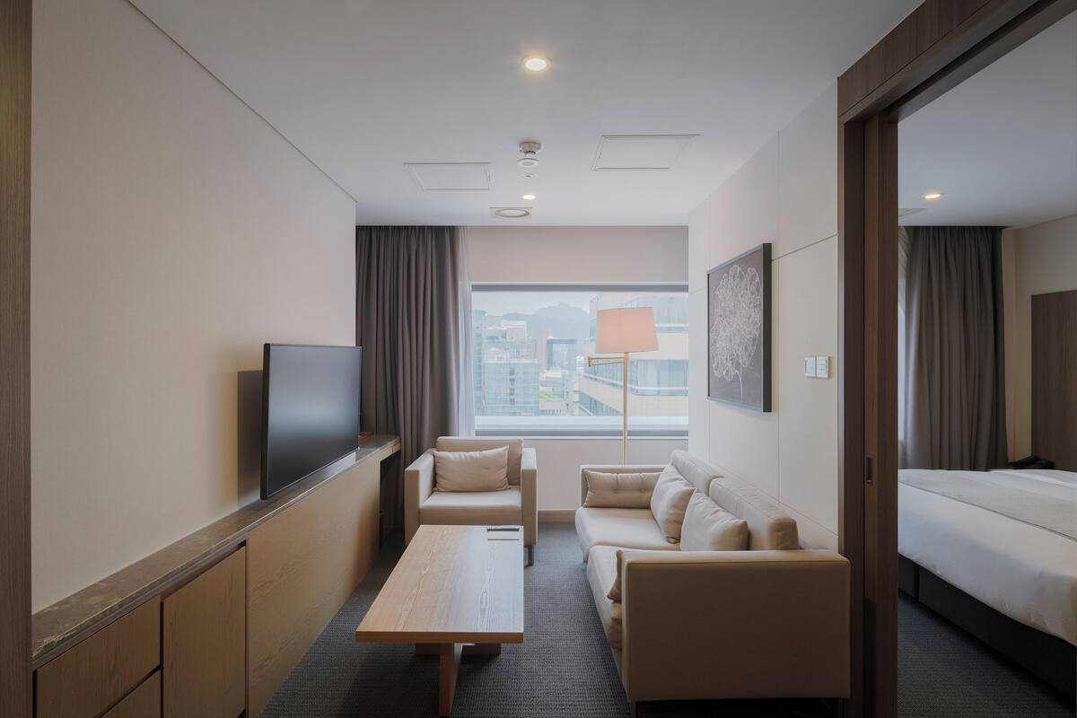 A hotel room with a flat-screen TV, a sofa, a coffee table, a bed, and a large window with a view of buildings.
