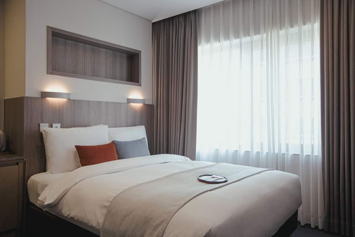 A hotel room with a large bed, a bedside table, a built-in shelf above the bed, and sheer curtains on the window.