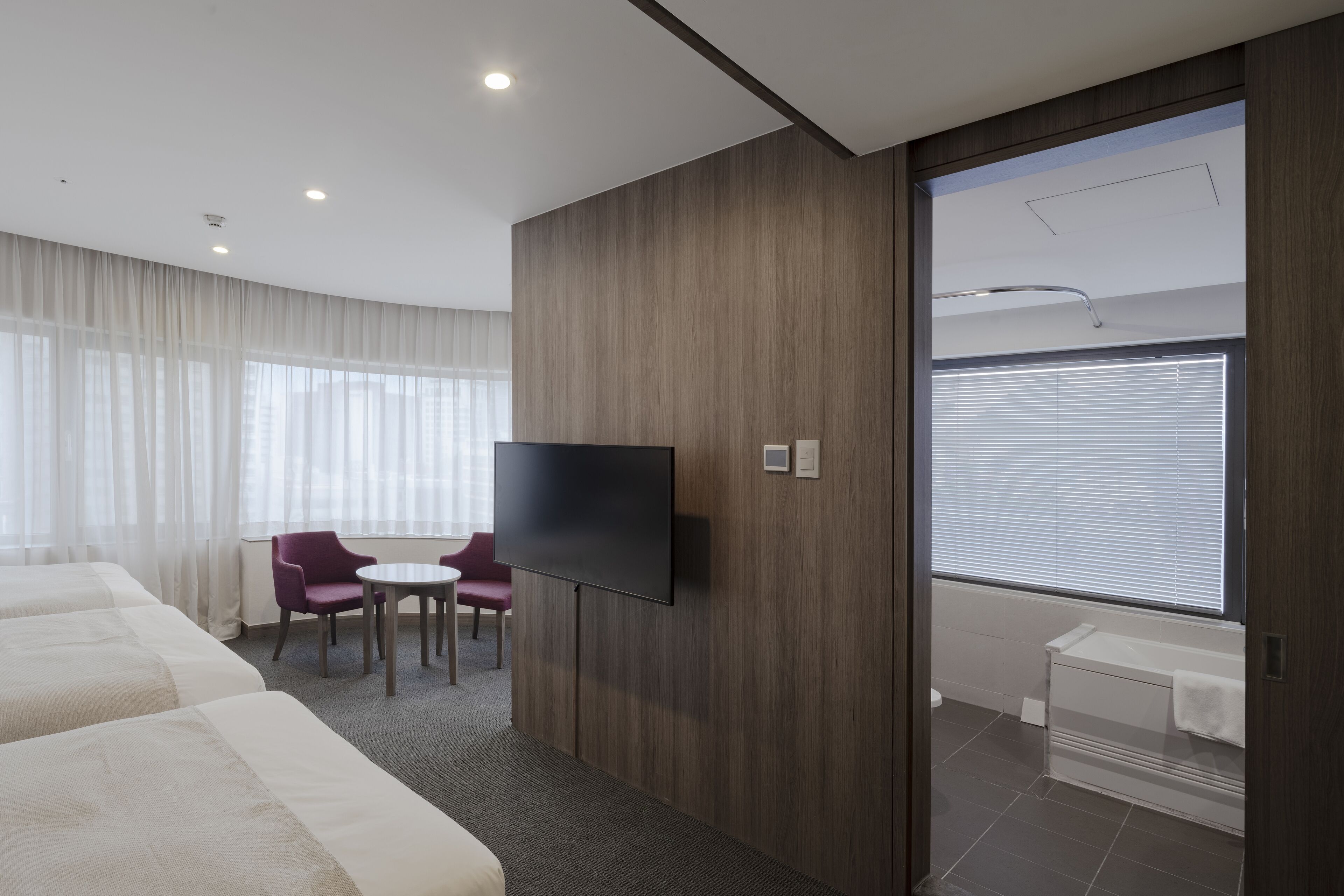 A hotel room with a flat-screen TV, a bed, a desk, a chair, and a window with blinds.