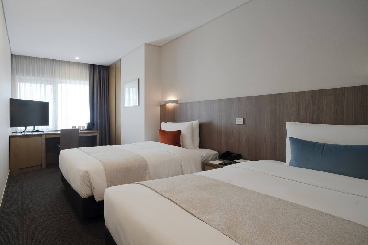 A hotel room with two beds, a desk, a TV, and a window with curtains.