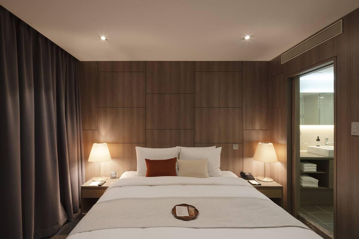 A hotel room with a large bed, two bedside tables with lamps, a nightstand with a phone, and a view of the bathroom through a glass door.