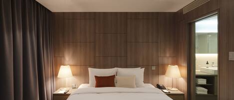 A hotel room with a large bed, two bedside tables with lamps, a nightstand with a phone, and a view of the bathroom through a glass door.