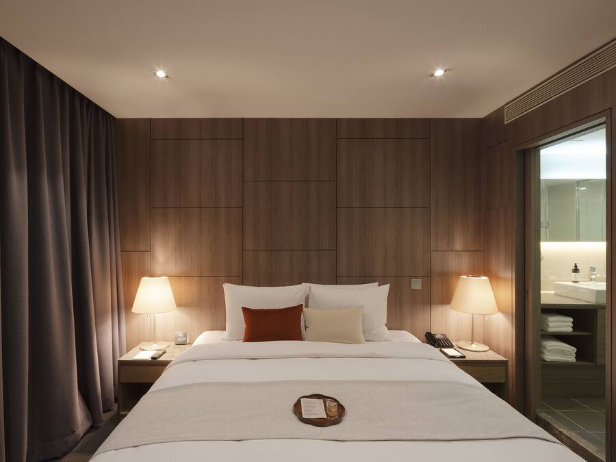 A hotel room with a large bed, two bedside tables with lamps, a nightstand with a phone, and a view of the bathroom through a glass door.