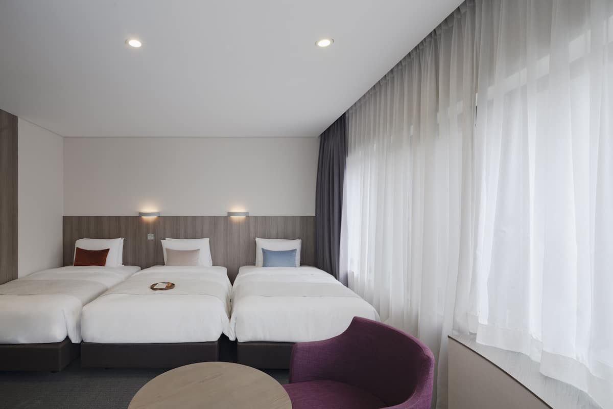 A hotel room with two beds, a small round table, a purple chair, and a large window with sheer curtains.