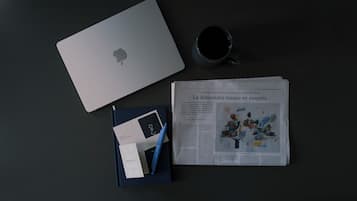 Una computadora portátil, una taza, un cuaderno y un periódico sobre una superficie oscura.