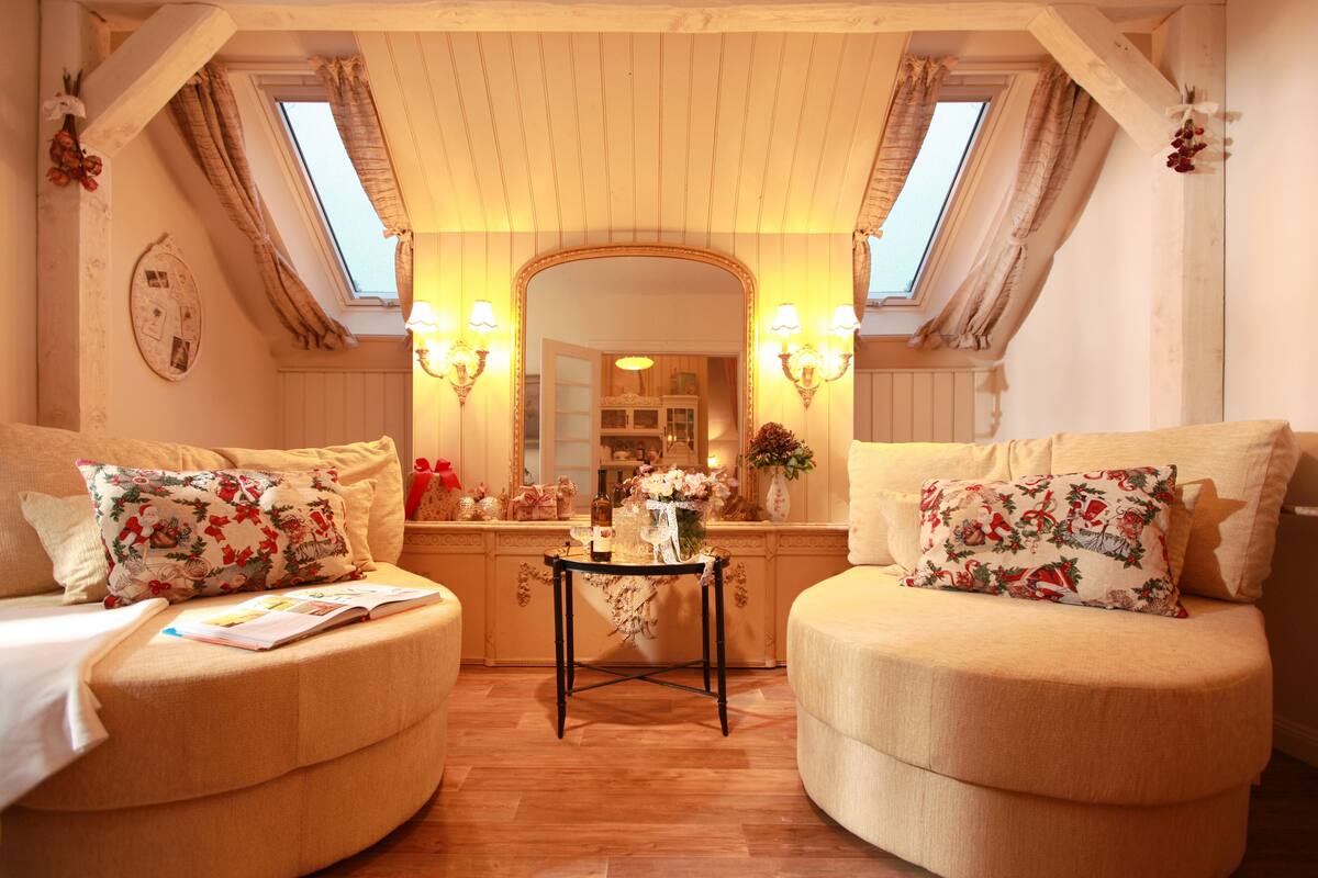 A cozy room with two armchairs, a small table, and a mirror. There are two windows with curtains, a skylight, and a wall-mounted light fixture.