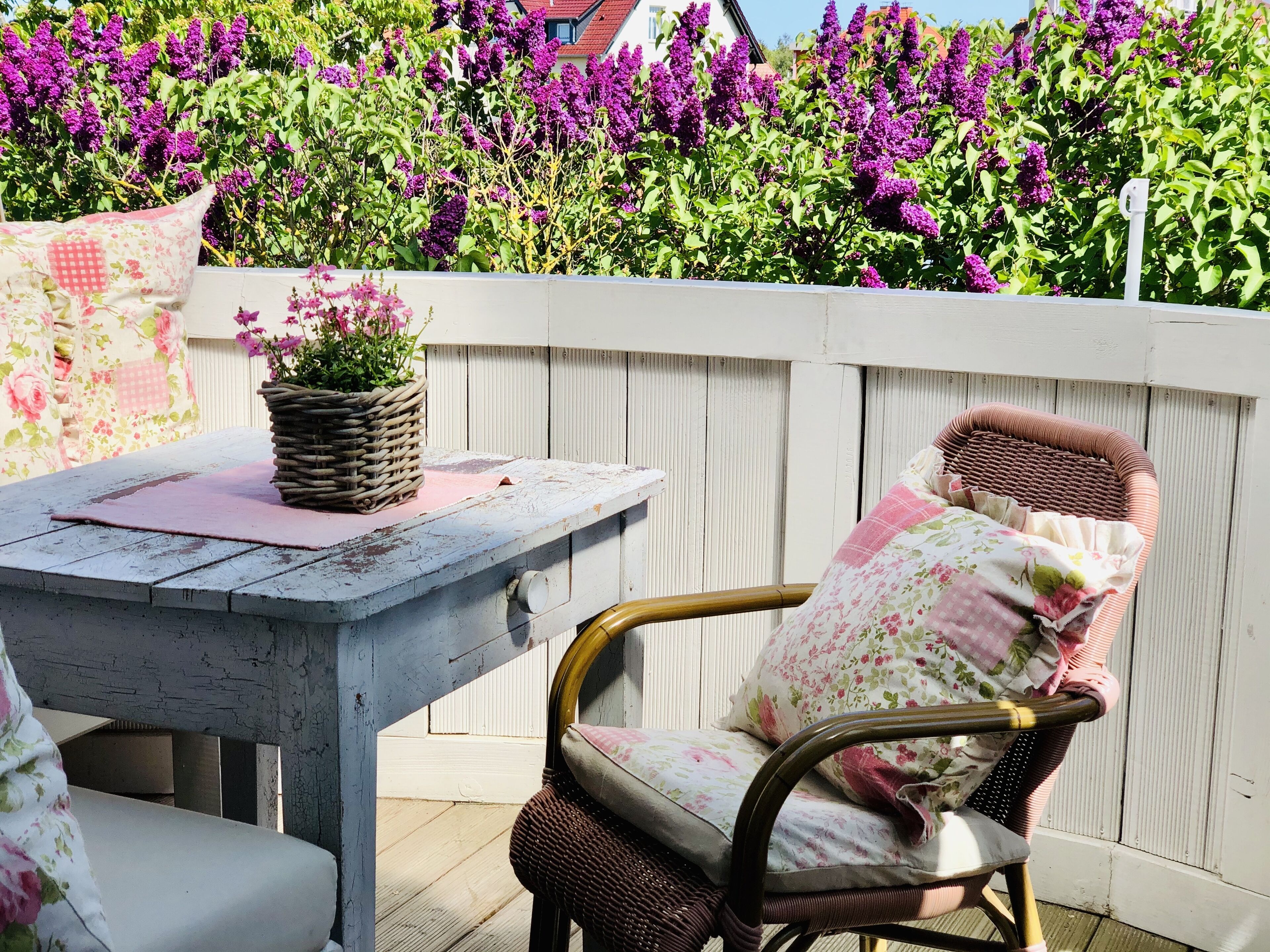 Eine Terrasse mit Tisch und Korbstühlen, umgeben von einem weißen Zaun und lila Blumen.