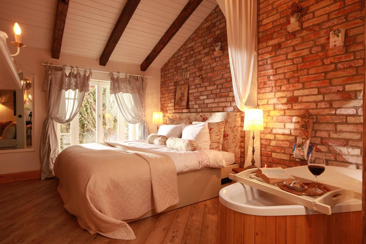 A cozy bedroom with a brick wall, wooden beams, a large bed, a bedside table with a wine glass, and a window with curtains.