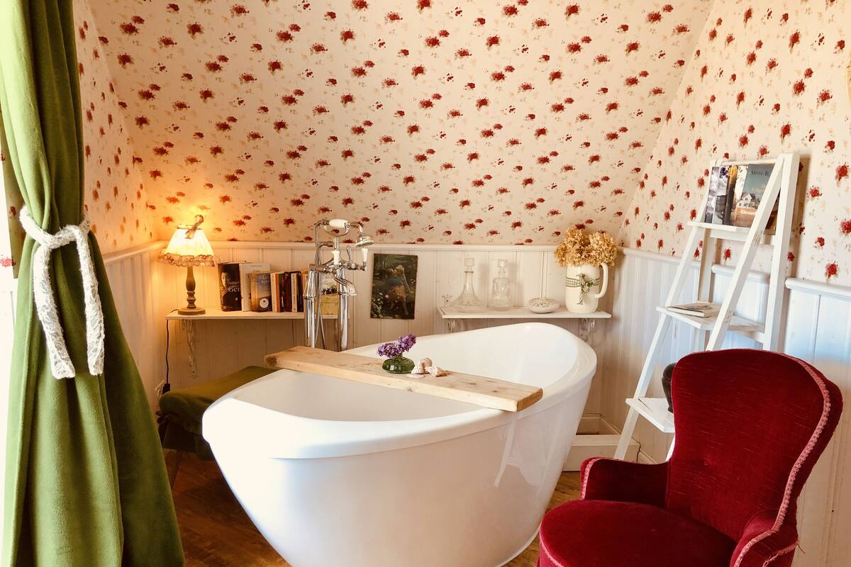 A bathroom with a freestanding bathtub, a red chair, and a floral wallpaper pattern.