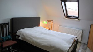 Un dormitorio con cama, una silla, una mesita de noche con lámpara, una ventana y un radiador.