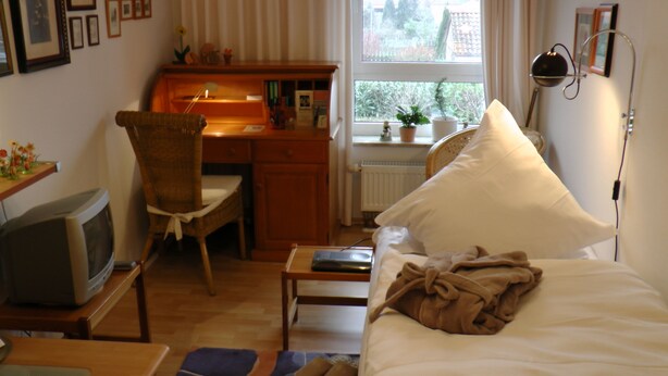 Un dormitorio con cama, escritorio, silla, televisor y ventana con vista a árboles.