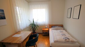 Una habitación pequeña y luminosa con un escritorio de madera, una silla, una cama con colcha estampada, una planta en maceta y dos cuadros en la pared.
