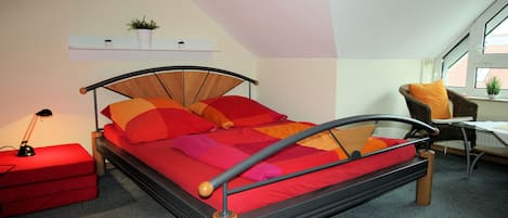 A bedroom with a bed, a nightstand, a chair, a table, and a shelf with a plant.