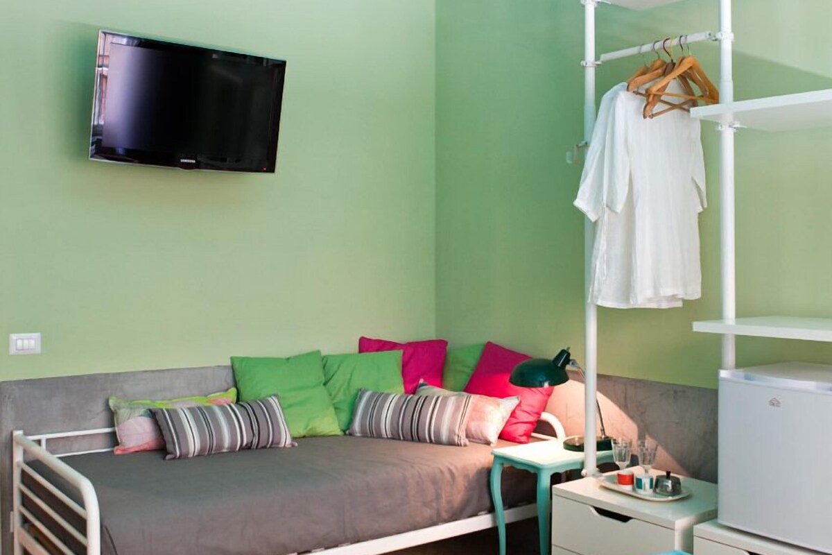 A room with a bed, a TV mounted on the wall, a hanging rack with clothes, and a blue exercise ball.