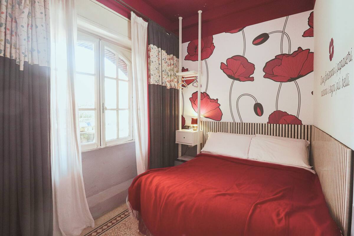 A bedroom with a bed, curtains, a nightstand, a lamp, and a wall with floral wallpaper.