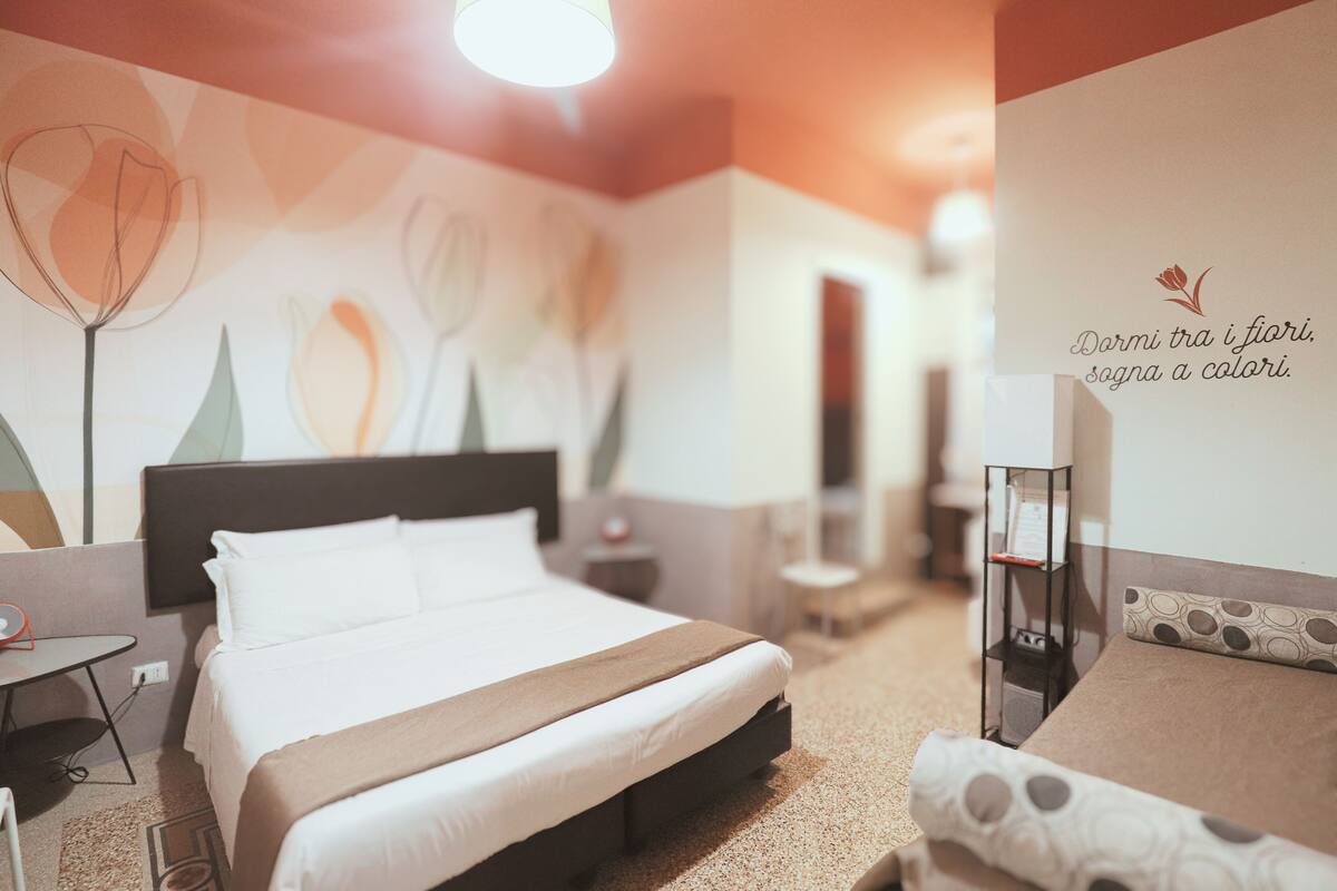 A hotel room with a bed, a bedside table, a chair, a desk, and a wall with a floral pattern.