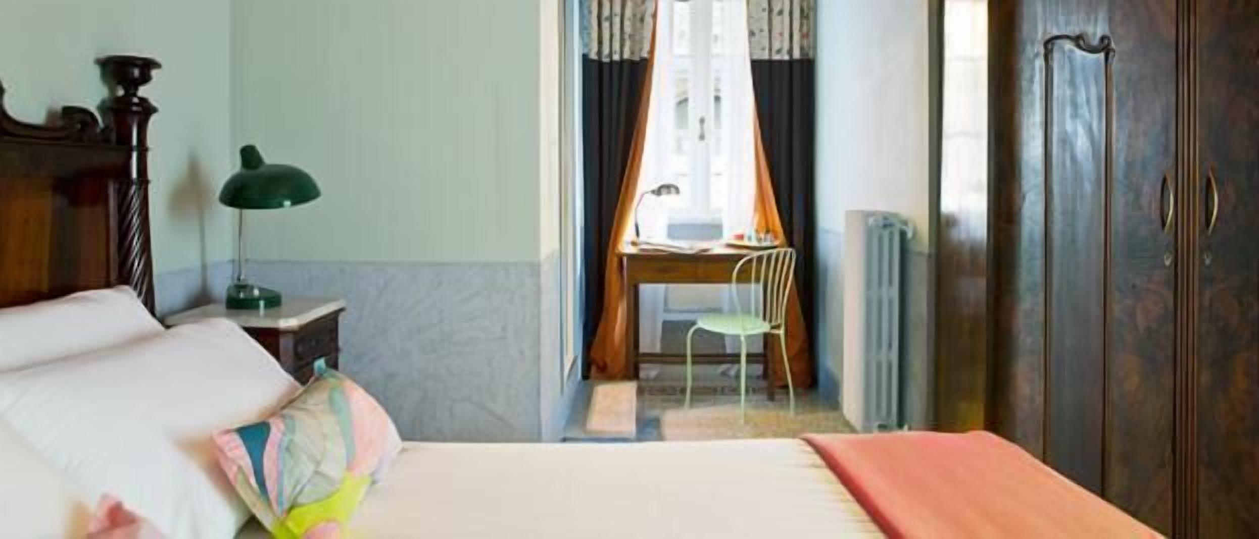 A hotel room with a bed, a desk with a chair, a lamp, and a wardrobe.