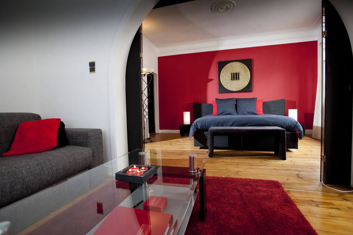 Un dormitorio moderno con una pared de acento roja, una cama grande, un sofá y una mesa de centro de vidrio.