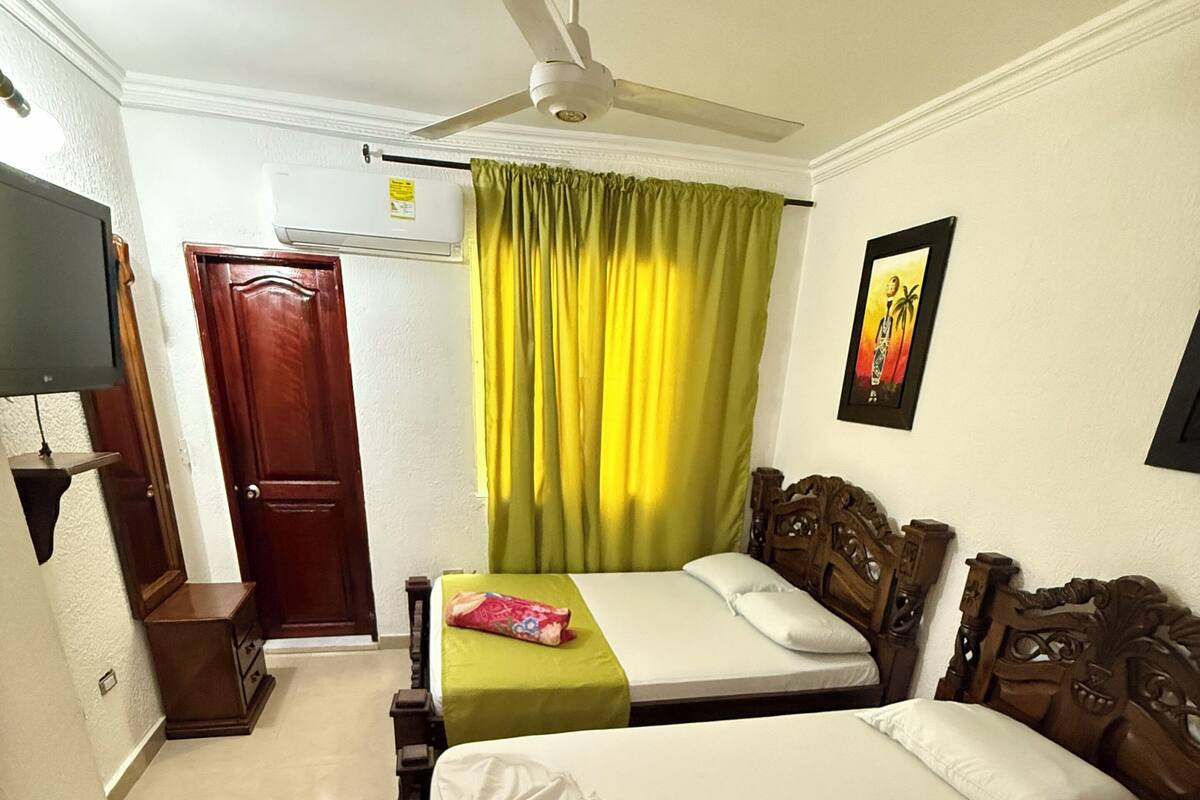 A hotel room with a bed, a ceiling fan, a green curtain, a wooden headboard, and a framed picture on the wall.