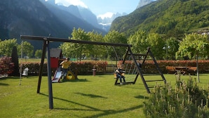 A playground with swings and slides, surrounded by a well-maintained lawn and flowering bushes, with mountains in the background. - Garnì Lago Alpino (Molveno)