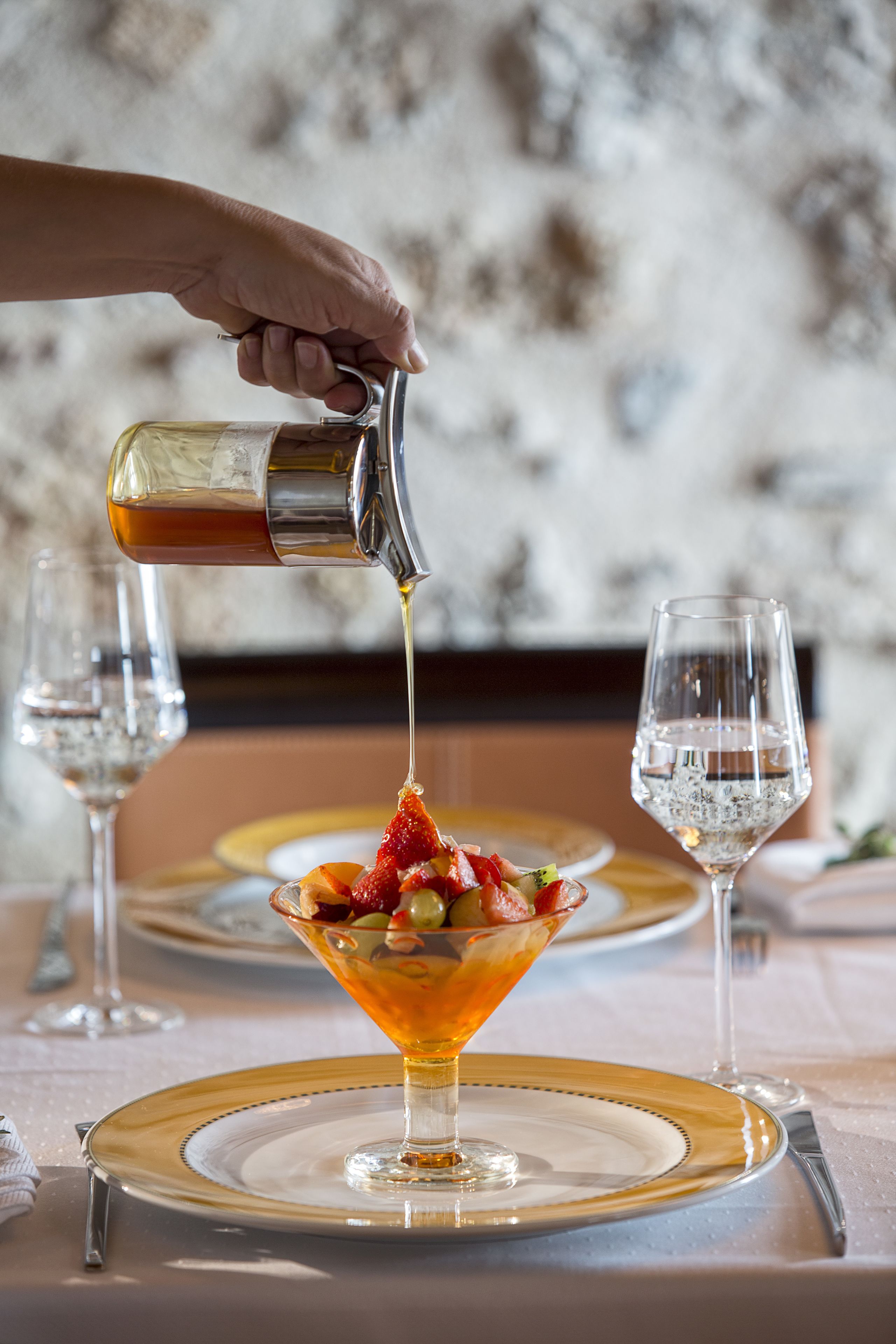Una mano sirve una bebida en un vaso sobre una mesa con un plato de borde dorado y un vaso transparente.
