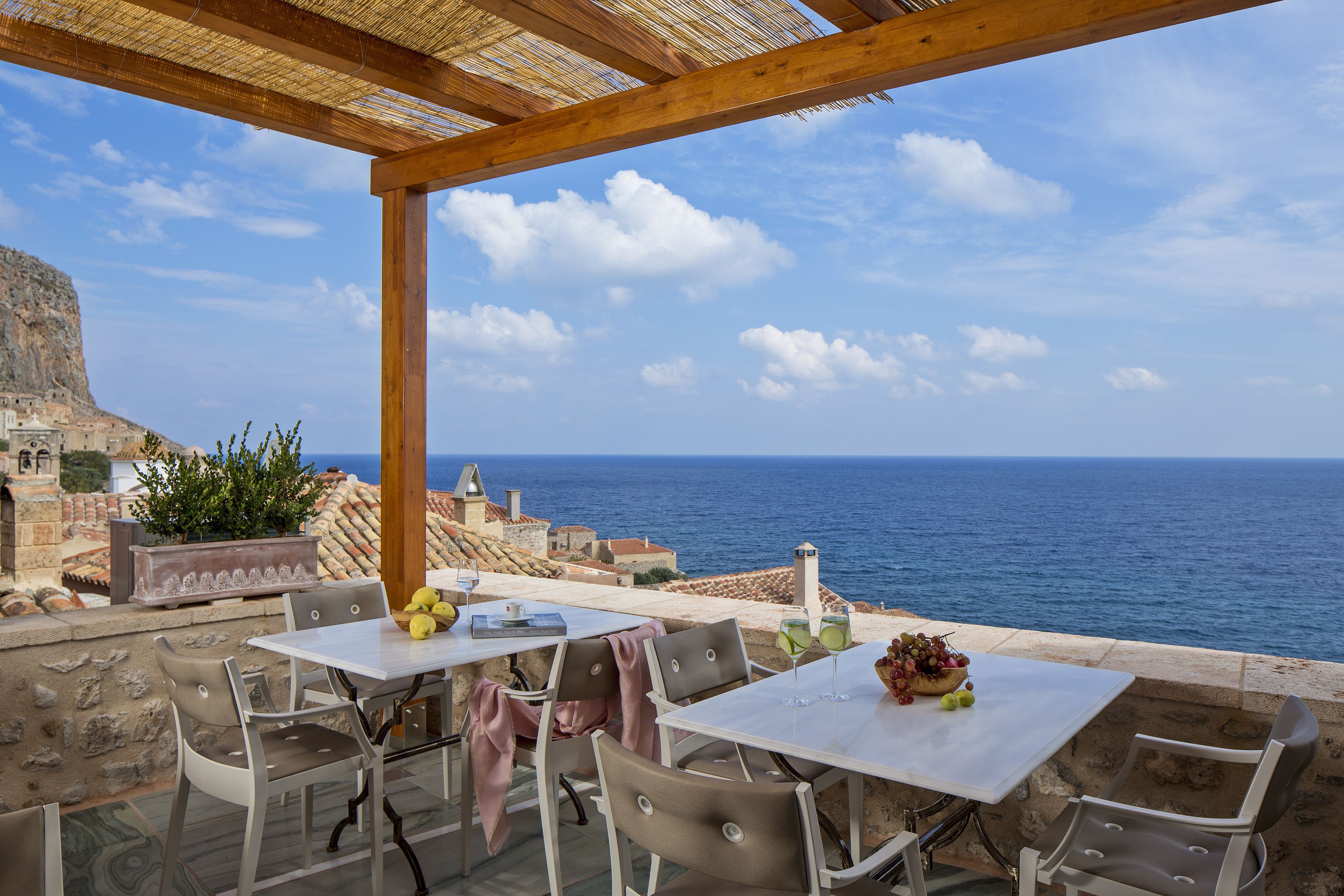 An outdoor seating area with tables and chairs overlooking the sea, featuring a stone wall and a view of the coastline.
