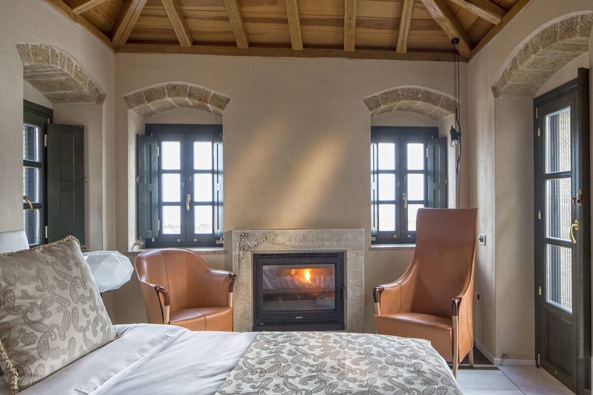 Una habitación con chimenea, dos sillones de cuero, una cama con ropa de cama estampada y arcos de piedra.