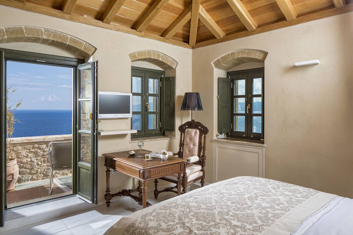 Un dormitorio con cama, escritorio, silla y televisor. Se ve el mar a través de una puerta abierta.