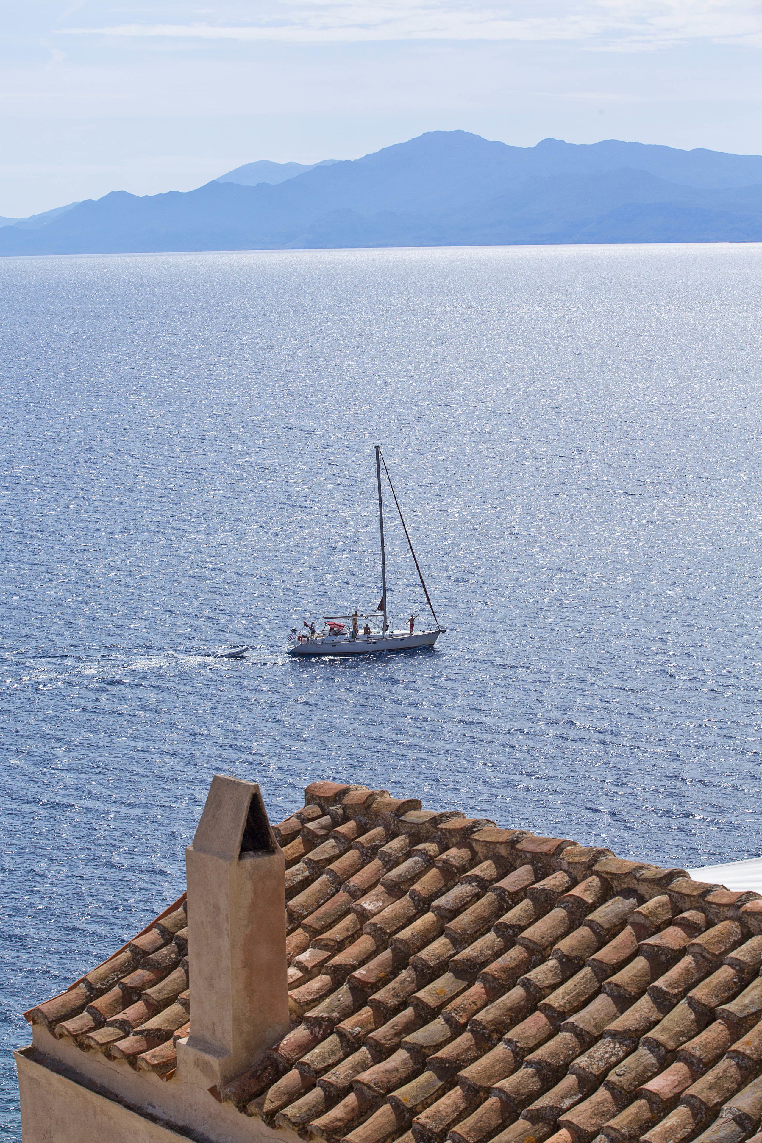 A coastal scene with a sailboat on the water and a tiled roof in the foreground.