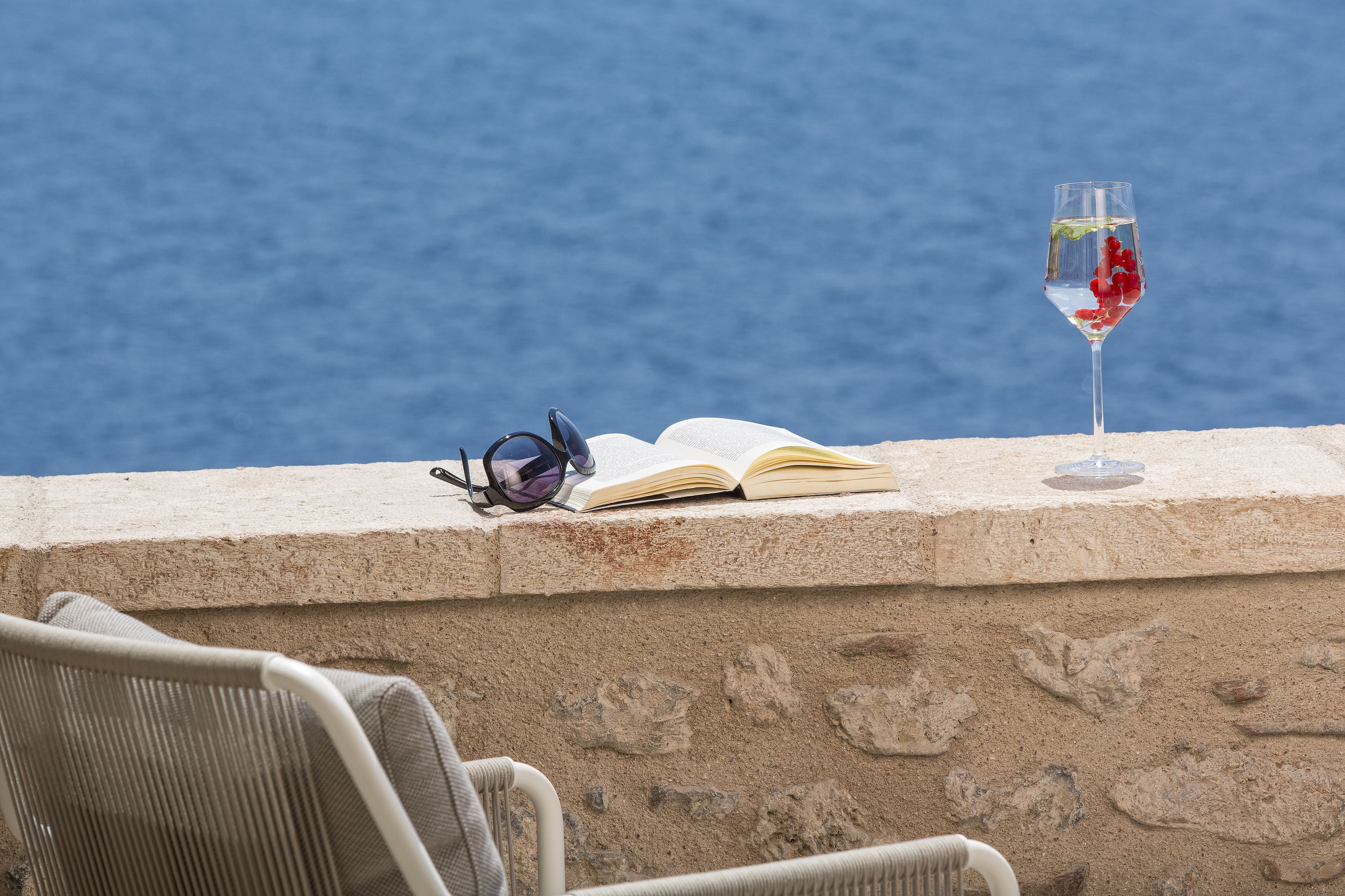 Un libro abierto y una copa de vino tinto sobre un borde con vista al mar.