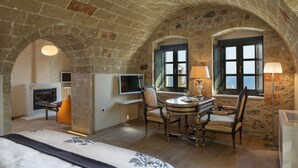 A room with a stone ceiling, a fireplace, a television, a dining table with chairs, and two windows with shutters. - Moni Emvasis Luxury Suites (Monemvasia)