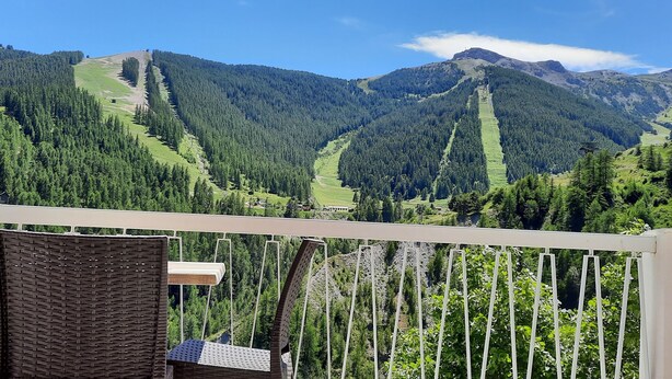 A balcony with wicker chairs and a view of a mountainous landscape with dense forests and a clear sky.
