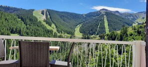 A balcony with wicker chairs and a view of a mountainous landscape with dense forests and a clear sky.