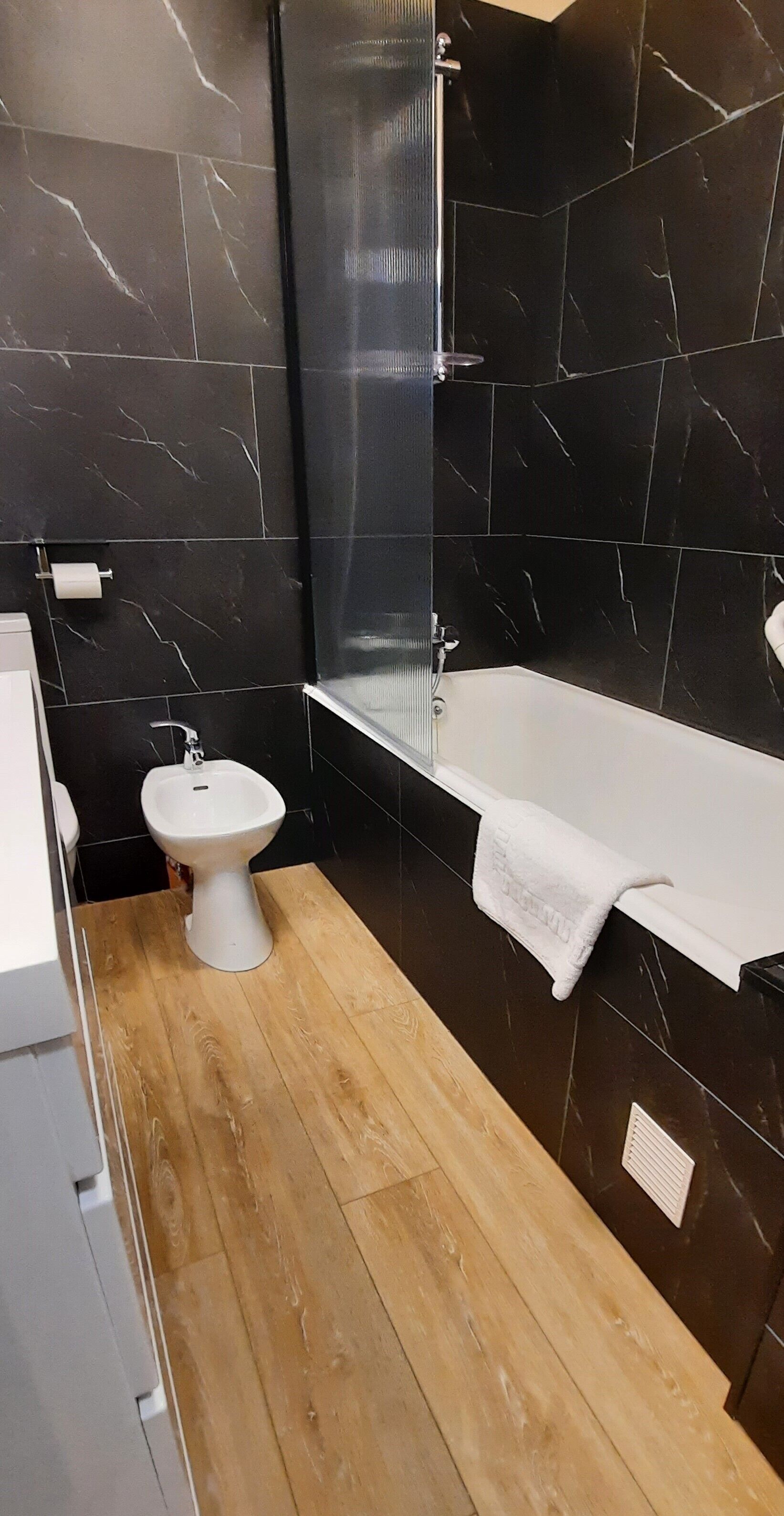 Un baño moderno con bañera, lavamanos pedestal blanco y un bidé. Las paredes están revestidas con baldosas de mármol negro, y el piso es de madera.