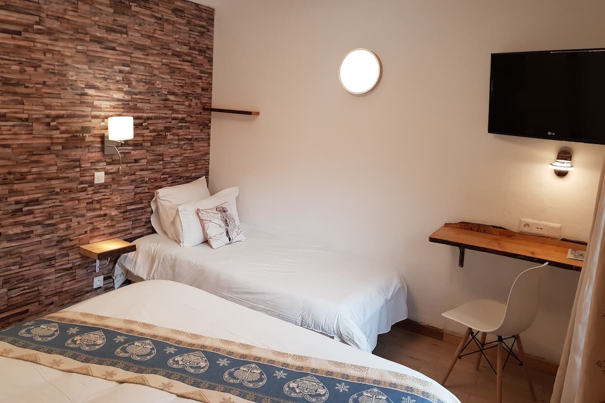 A bedroom with a wooden wall, a bed with patterned bedding, a bedside table, a desk with a chair, and a TV mounted on the wall.