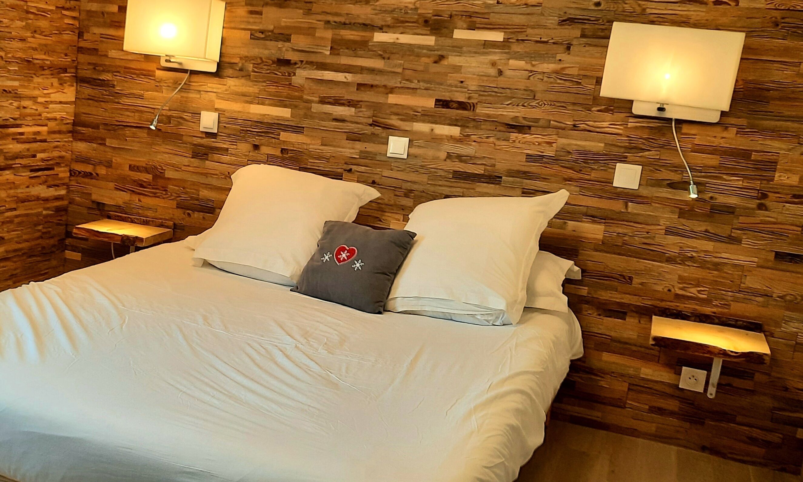 A neatly made bed with white linens and two pillows, a gray pillow with a heart and snowflake design, and two bedside lamps on wooden walls.