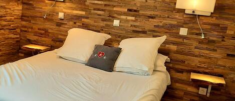 A neatly made bed with white linens and two pillows, a gray pillow with a heart and snowflake design, and two bedside lamps on wooden walls.