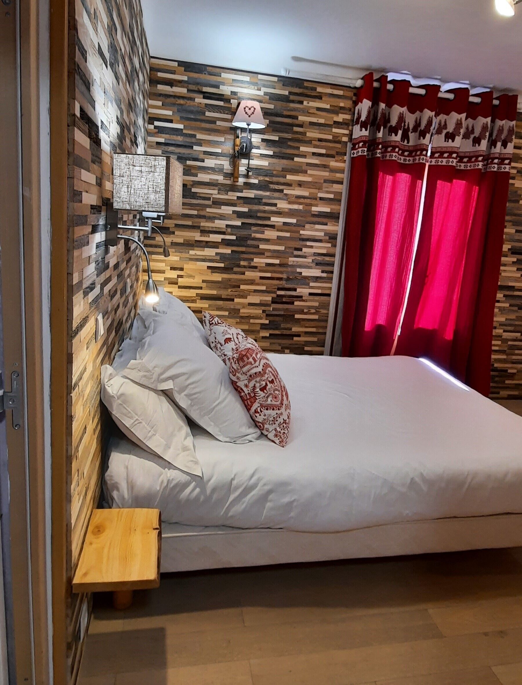 Une chambre avec un mur en bois, un lit avec une literie blanche, un coussin rouge à motifs, une table de chevet et une lampe fixée au mur.