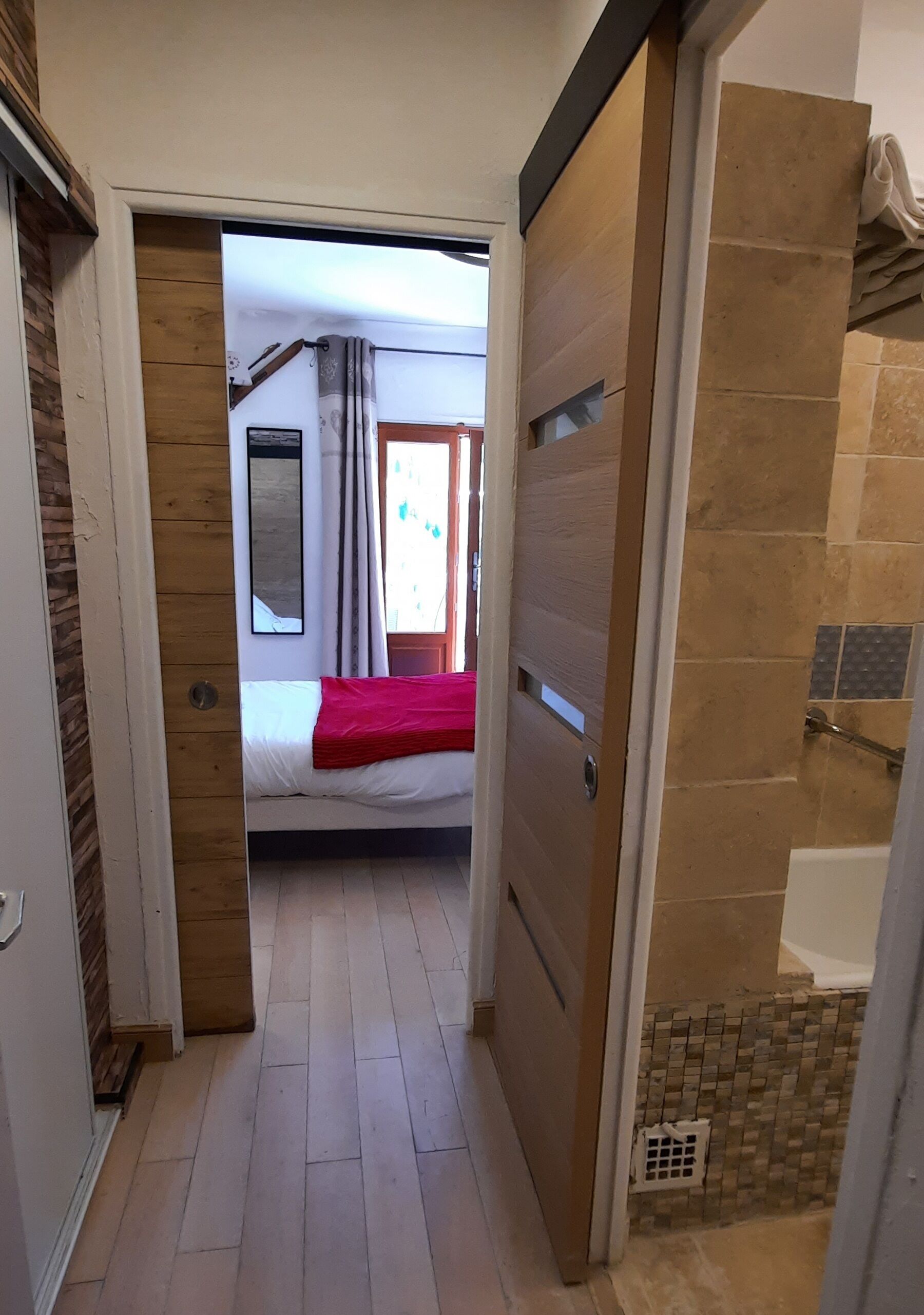 A narrow hallway leading to a bedroom with a bed, a red blanket, and a window with curtains.