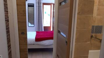 A narrow hallway leading to a bedroom with a bed, a red blanket, and a window with curtains.