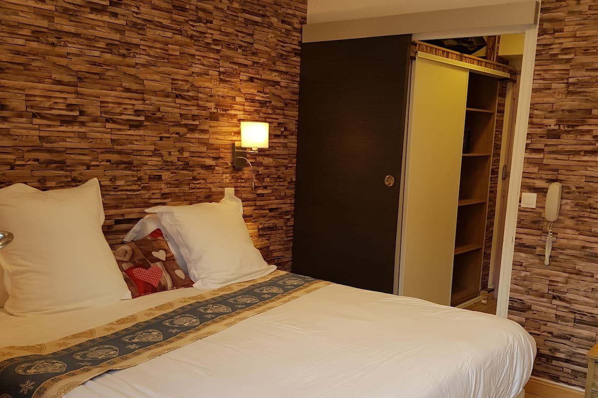 A neatly made bed with white linens and a decorative throw, a wooden wall with a built-in shelf, and a bedside telephone.
