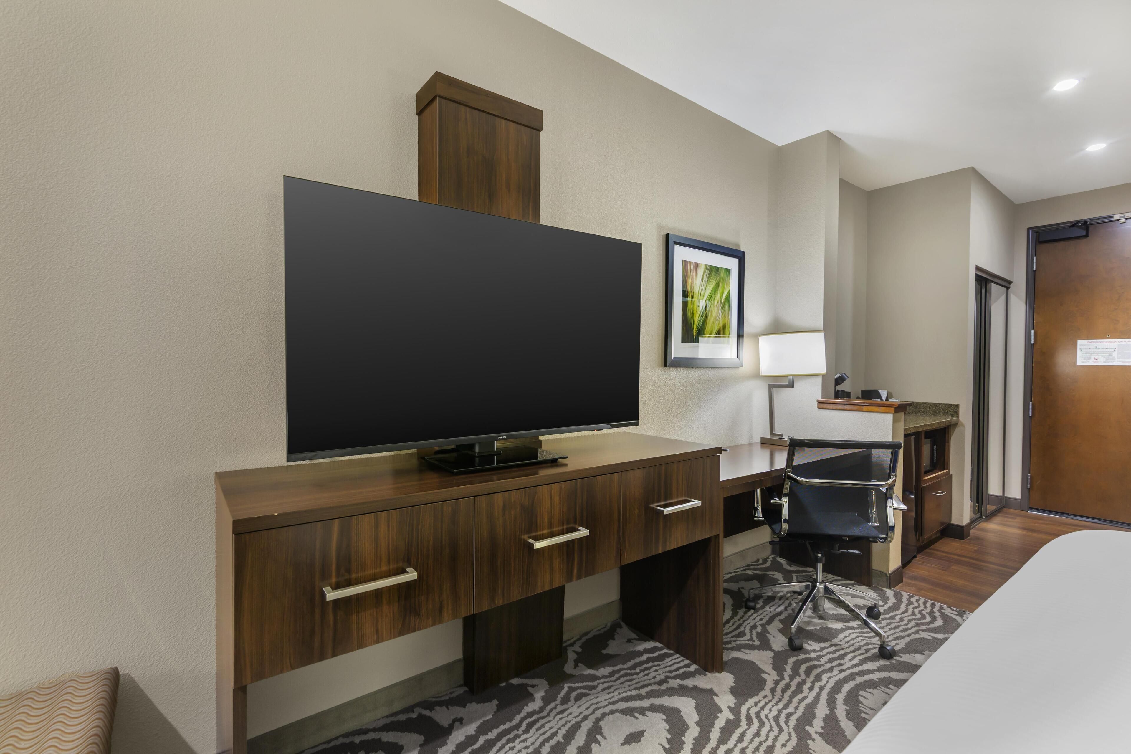 A hotel room with a large flat-screen TV on a wooden desk, a chair, a bed, and a door.