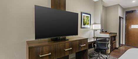 A hotel room with a large flat-screen TV on a wooden desk, a chair, a bed, and a door.