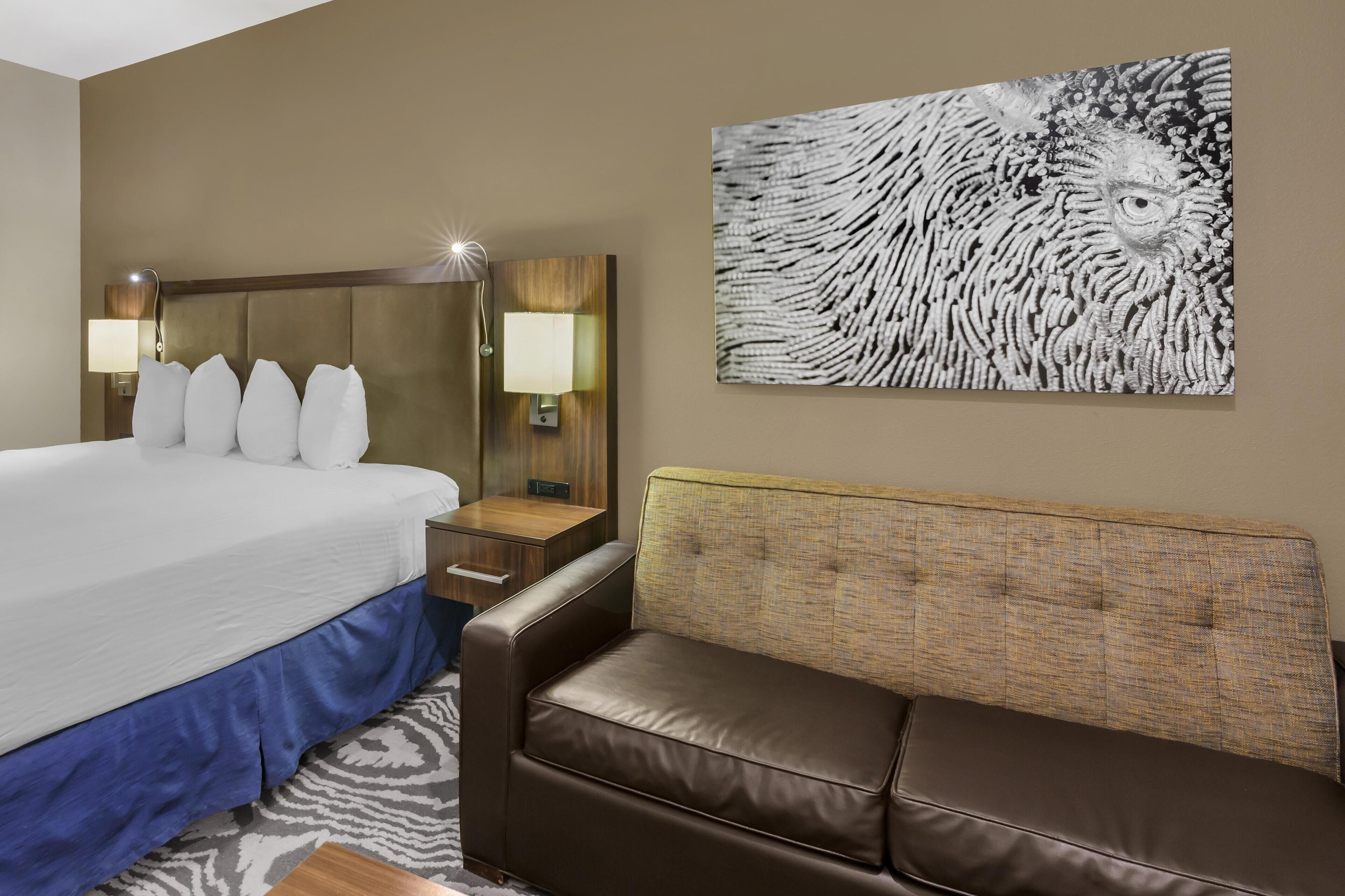 Una habitación de hotel con cama, sofá de cuero marrón, lámpara de pared y un cuadro abstracto grande.