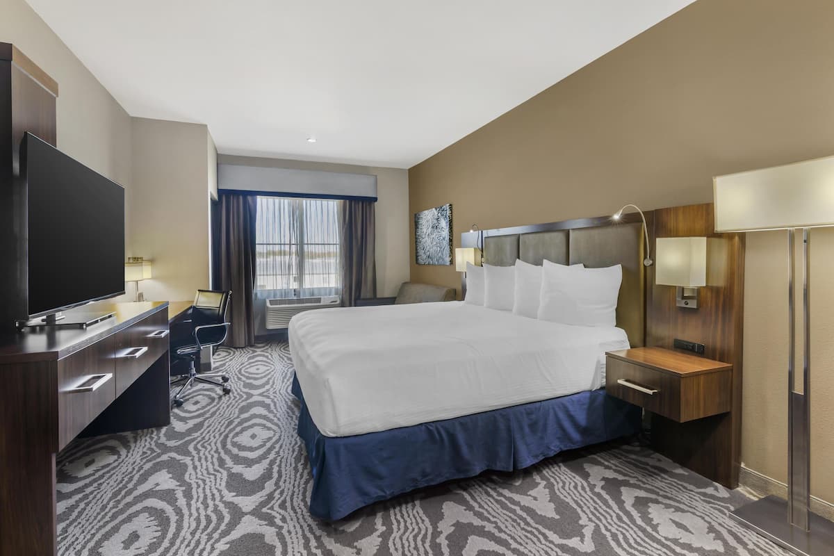 Quarto de hotel com uma cama grande, uma escrivaninha com televisão de tela plana, uma cadeira e uma janela com cortinas.