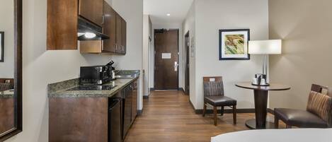 A hotel room with a kitchenette, a bed, a seating area with a table and chairs, and a hallway leading to the door.